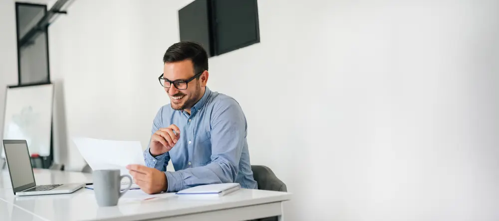 Lachender Mann sitzt am Schreibtisch vor dem Laptop.